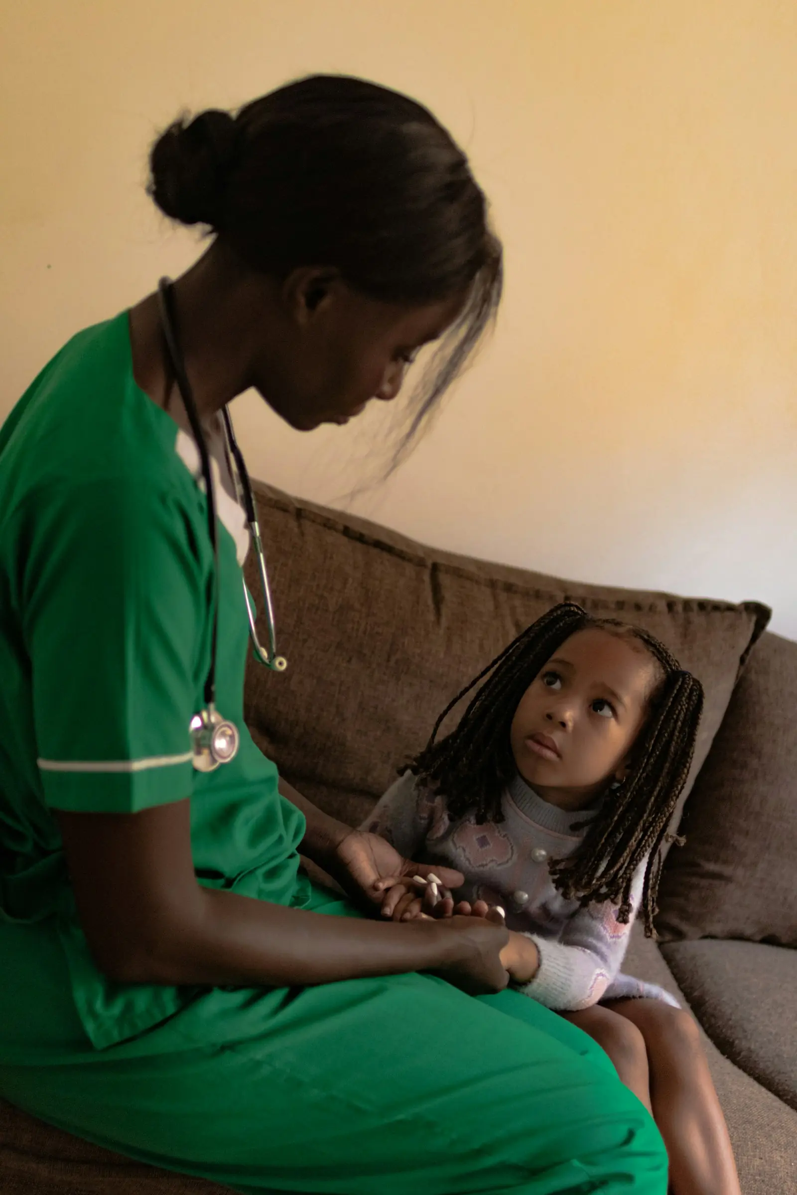 A nurse in green scrubs sits with a young girl on a sofa at home, holding her hands.