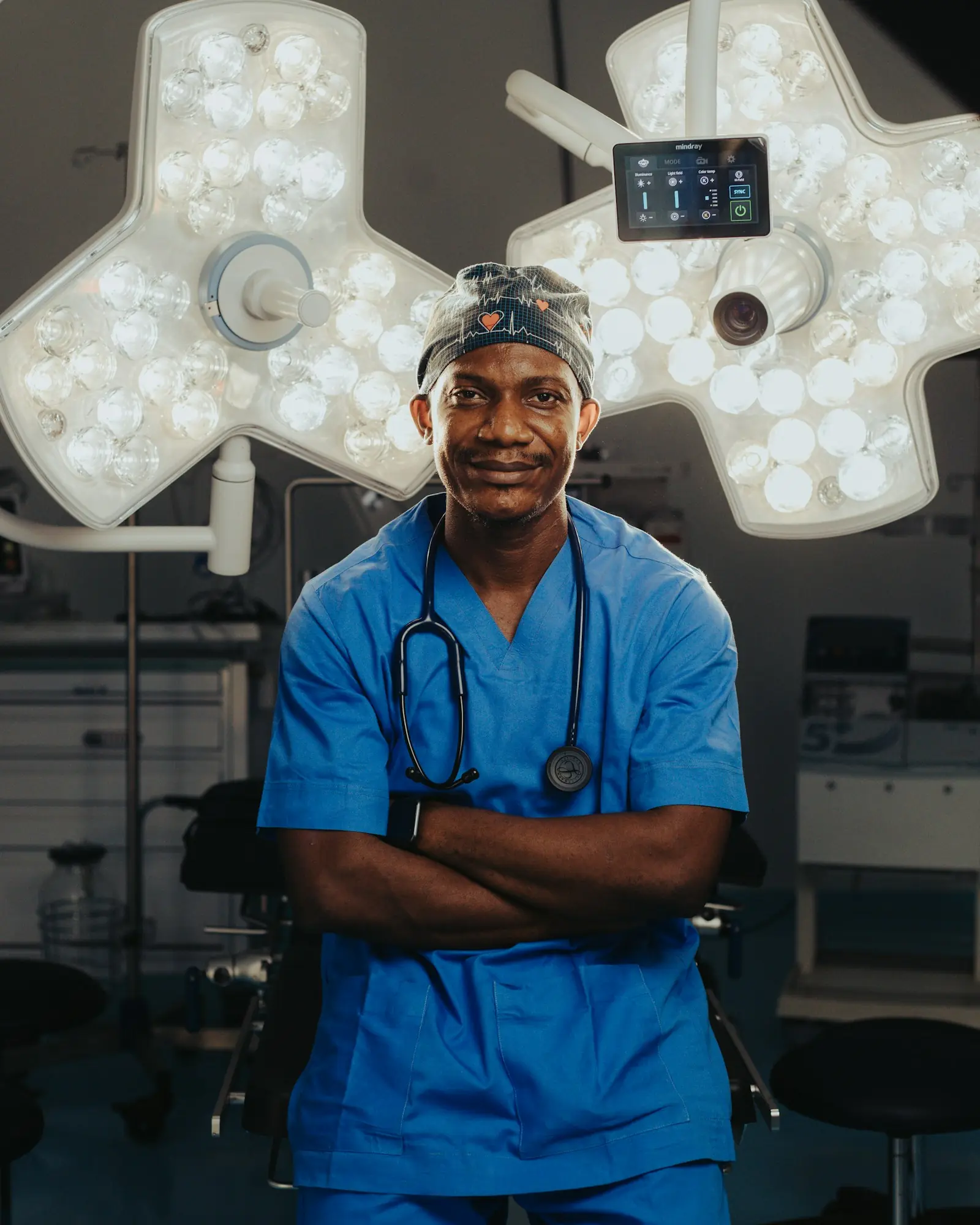A Nigerian doctor in blue scrubs with a stethoscope, arms folded, smiling.